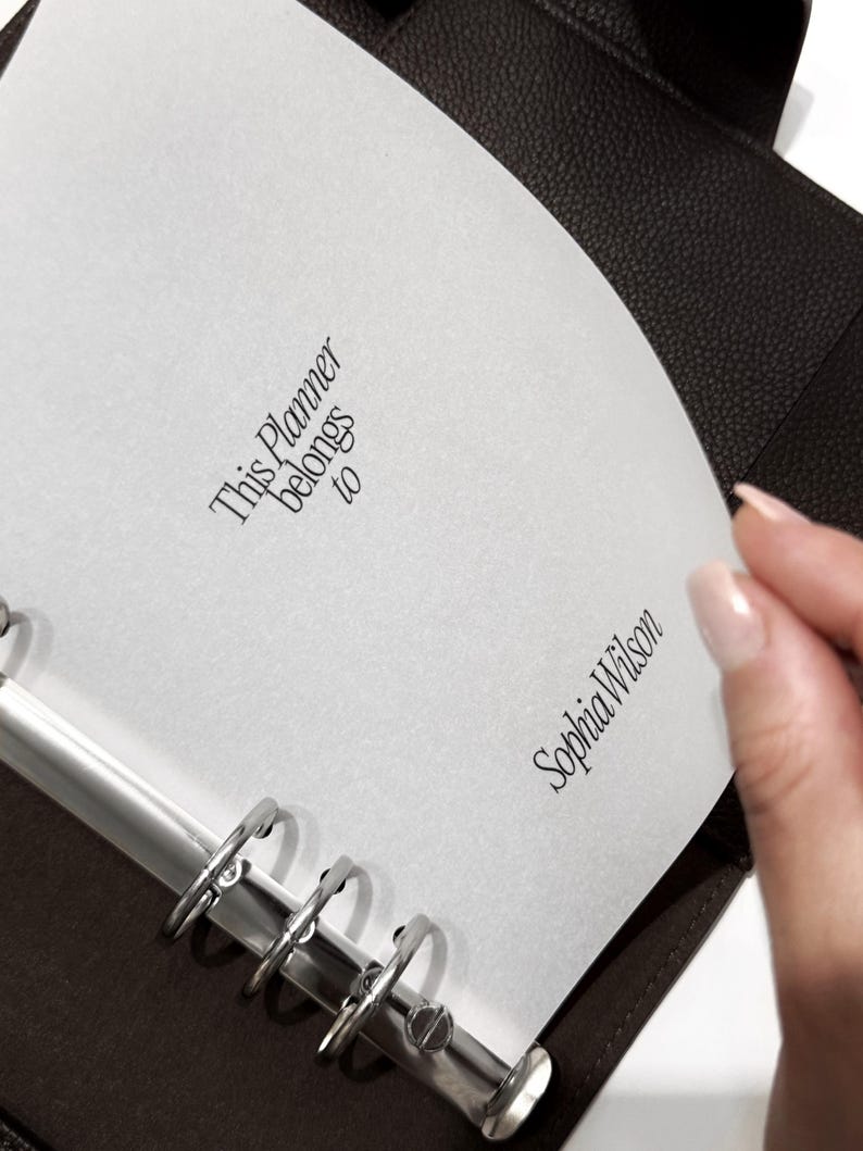 May include: A dark brown leather planner with silver metal rings. The open page reads "This Planner belongs to" and "Sophia Wilson" in elegant script. The planner is partially open, revealing the interior pages.