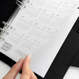 May include: A black leather ring-bound planner featuring a white calendar insert for the year twenty twenty seven. The page displays twelve months with numerical dates and the text twenty twenty seven printed in a serif font on the bottom left.