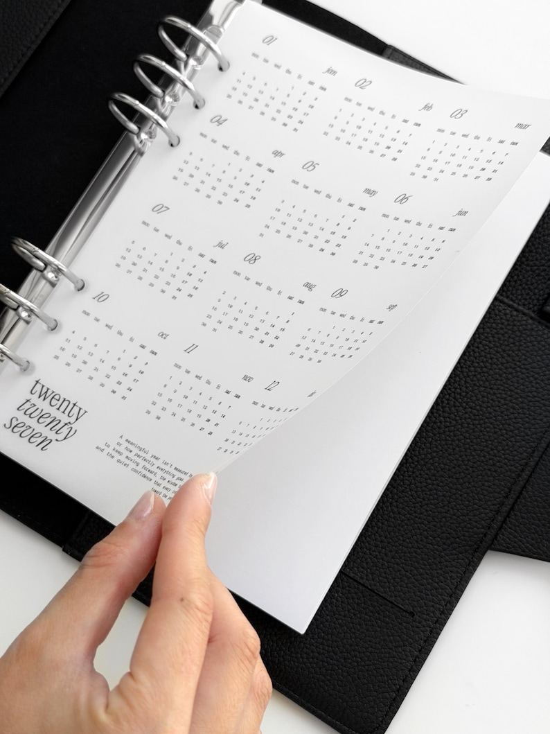 May include: A black leather ring-bound planner featuring a white calendar insert for the year twenty twenty seven. The page displays twelve months with numerical dates and the text twenty twenty seven printed in a serif font on the bottom left.