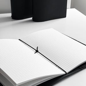May include: An open black notebook with grid paper, held closed by a black elastic band. The notebook is on a white surface, with two additional black notebooks in the background.