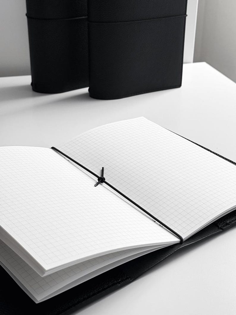 May include: An open black notebook with grid paper, held closed by a black elastic band. The notebook is on a white surface, with two additional black notebooks in the background.