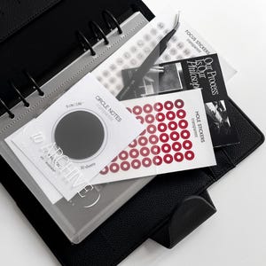 Op de afbeelding: Een zwarte leren binder met zilveren ringen, met diverse schrijfwaren. Inclusief stickers, transparante vellen en kaarten met tekst zoals "Circle Notes". De binder is open en toont de inhoud.