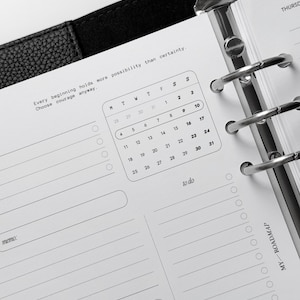 May include: A black leather planner with a silver six-ring binder mechanism. The white paper page features a monthly calendar, a to-do list, and a memo section. The text reads: Every beginning holds more possibility than certainty. Choose courage anyway.