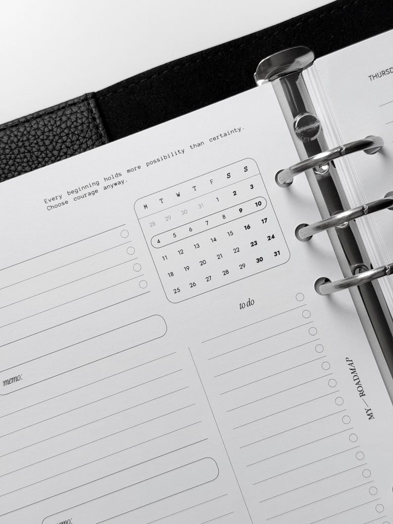 May include: A black leather planner with a silver six-ring binder mechanism. The white paper page features a monthly calendar, a to-do list, and a memo section. The text reads: Every beginning holds more possibility than certainty. Choose courage anyway.