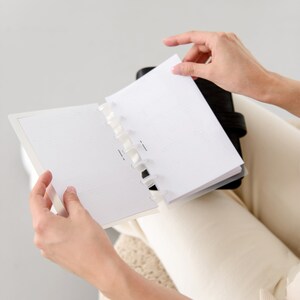 May include: A black leather-bound planner with dot grid pages is open, revealing a blank page. The planner is held by a person, with hands visible. The pages are white with a subtle dot pattern. The planner is open to a new page.