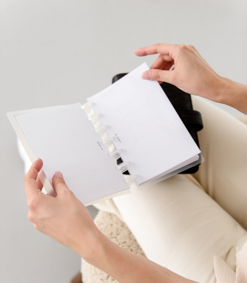 May include: A black leather-bound planner with dot grid pages is open, revealing a blank page. The planner is held by a person, with hands visible. The pages are white with a subtle dot pattern. The planner is open to a new page.