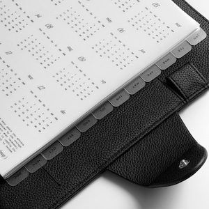 May include: A black leather-textured planner with a silver metal ring binder mechanism. The interior features white pages displaying a yearly calendar with months labeled from January to December, organized by tabbed dividers along the right edge.
