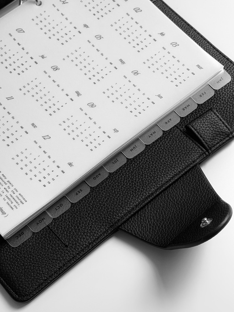May include: A black leather-textured planner with a silver metal ring binder mechanism. The interior features white pages displaying a yearly calendar with months labeled from January to December, organized by tabbed dividers along the right edge.