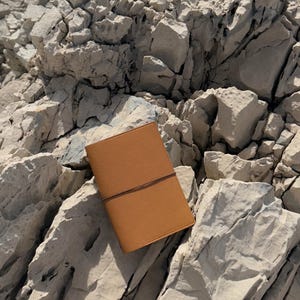 K&ouml;nnte beinhalten: Ein hellbraunes Lederjournal mit Riemenverschluss liegt auf hellgrauen Felsen. Das Journal hat eine strukturierte Oberfl&auml;che und ein schlichtes Design. Die Felsen sind rau und uneben und bilden eine nat&uuml;rliche Kulisse.