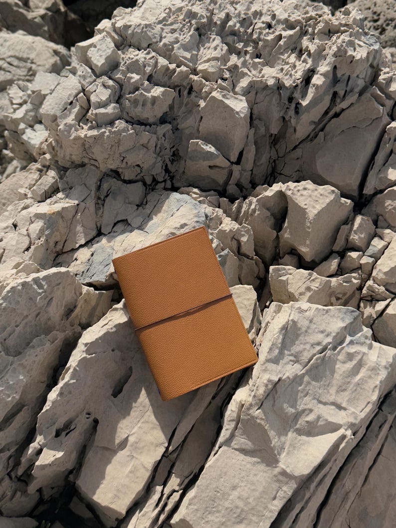K&ouml;nnte beinhalten: Ein hellbraunes Lederjournal mit Riemenverschluss liegt auf hellgrauen Felsen. Das Journal hat eine strukturierte Oberfl&auml;che und ein schlichtes Design. Die Felsen sind rau und uneben und bilden eine nat&uuml;rliche Kulisse.