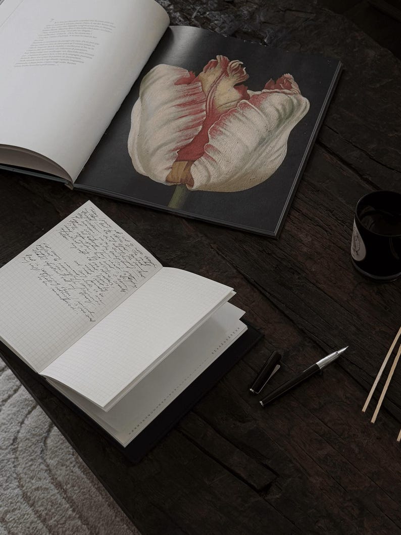May include: An open notebook with lined pages and handwritten text sits next to a pen and a book with a tulip illustration. The items are arranged on a dark wooden surface, with a cup and some sticks in the background.