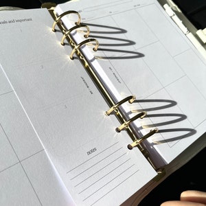 May include: Open planner with gold-colored rings and white pages. The planner is open to a page with a calendar layout and the word "notes" at the bottom. The planner is in a light-colored cover.