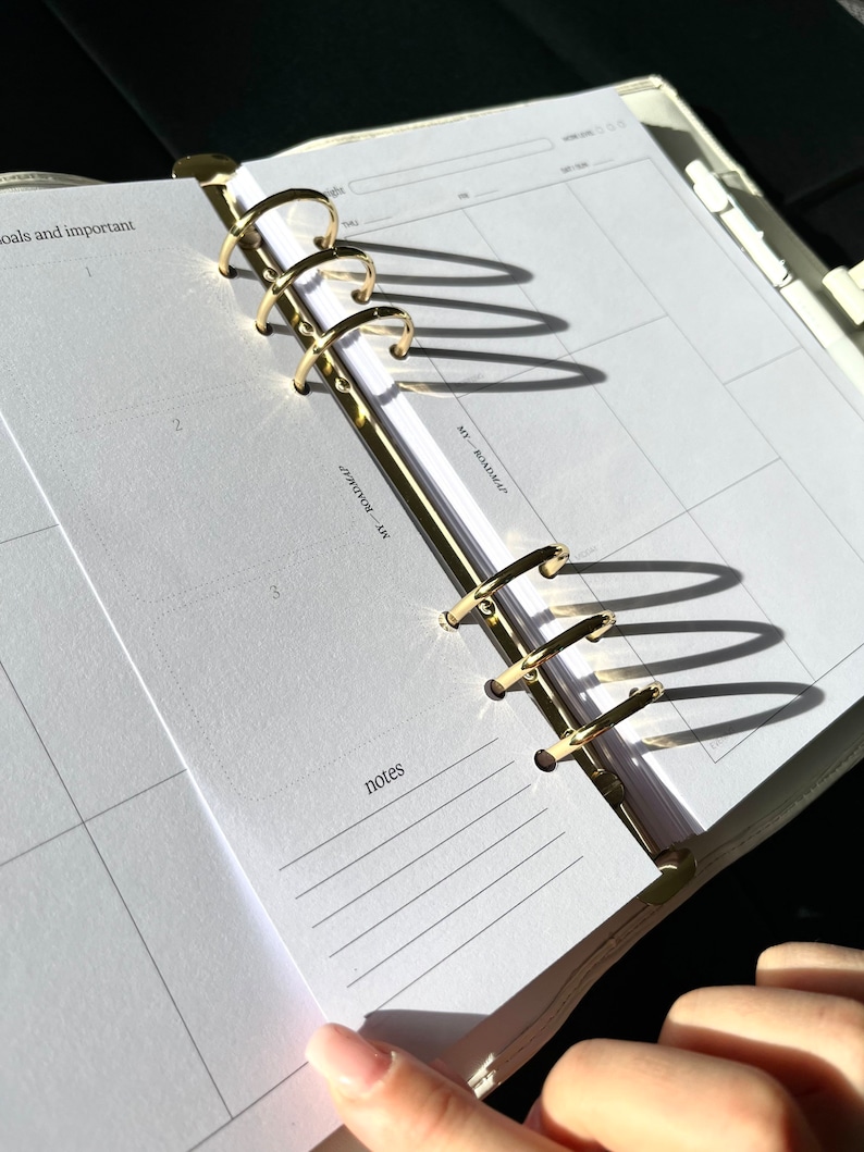 May include: Open planner with gold-colored rings and white pages. The planner is open to a page with a calendar layout and the word "notes" at the bottom. The planner is in a light-colored cover.