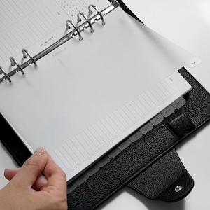 May include: A black textured leather binder with a six-ring metal mechanism holding white planner pages. The pages feature a monthly roadmap layout with a vertical list and a set of translucent tabbed dividers labeled with months from January to December.
