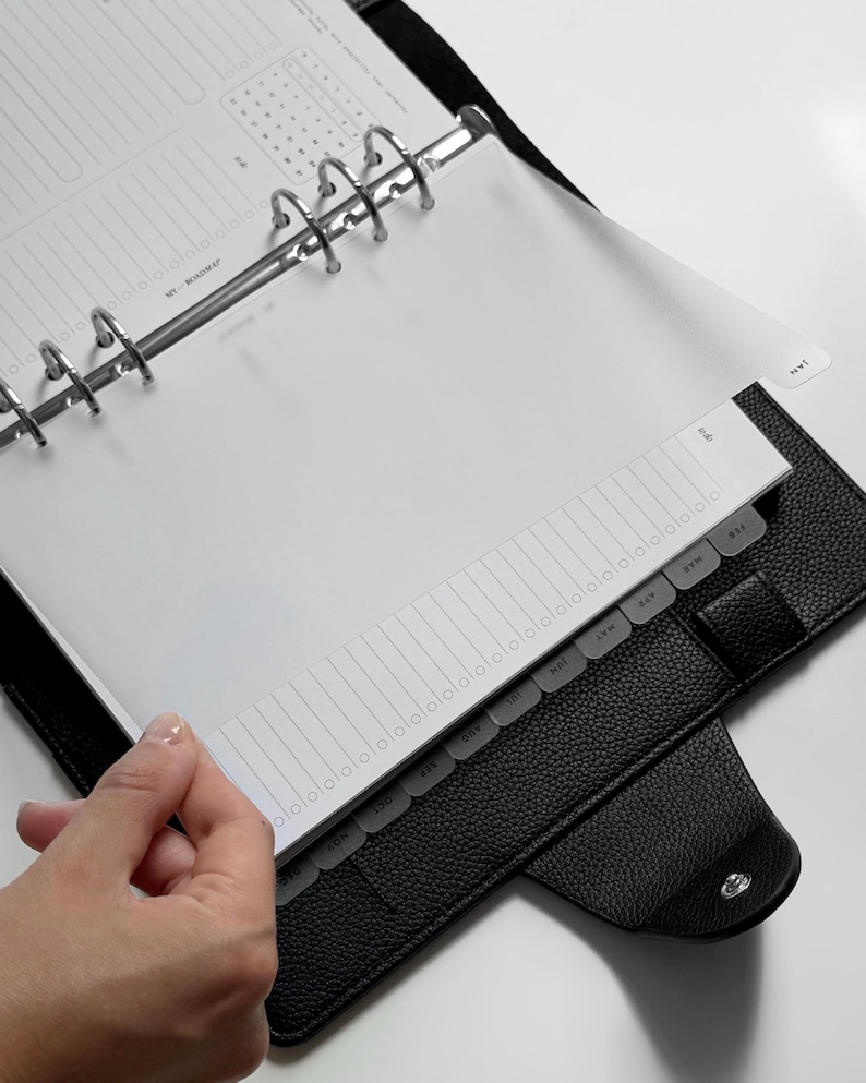 May include: A black textured leather binder with a six-ring metal mechanism holding white planner pages. The pages feature a monthly roadmap layout with a vertical list and a set of translucent tabbed dividers labeled with months from January to December.
