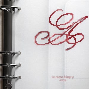 Può includere: Una pagina di agenda bianca con un raccoglitore ad anelli in metallo argentato sul lato sinistro. La pagina presenta un grande monogramma a punto croce rosso della lettera E e il testo stampato this planner belongs to Amelia in basso.