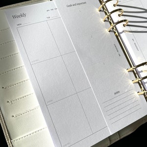 May include: A cream-colored planner with gold-colored rings, open to a weekly spread. The left page is labeled "Weekly" with spaces for each day. The right page is labeled "Goals and important" with numbered sections. The planner is on a black surface.