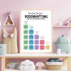 May include: A framed poster with the German text "5.4.3.2.1 BODENHAFTUNG" and instructions to identify things you see, touch, hear, smell, and taste. The poster has a white background with colorful illustrations of eyes, hands, ears, noses, and mouths.
