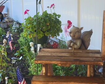May include: A small wooden picnic table with a squirrel figurine sitting on top. The table is in a garden setting with a white fence, flowers, and an American flag.