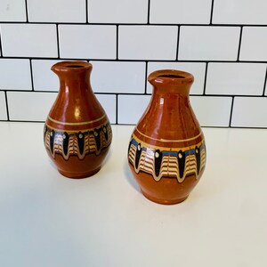 Pode incluir: Dois pequenos vasos de cor terracota com uma faixa decorativa de padrões geométricos pretos, brancos, azuis e amarelos. Os vasos têm um pescoço estreito e uma borda alargada, e são exibidos numa superfície branca.