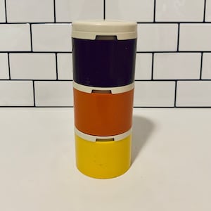 May include: A stackable container set with three sections in yellow, orange, and dark purple. Each section has a white lid and base. The containers are cylindrical and appear to be made of plastic.