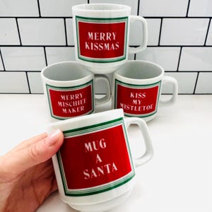May include: White ceramic mugs with red rectangular labels and green borders. Each mug has a different holiday-themed message in white text: "MERRY KISS-MAS", "MERRY MISCHIEF MAKER", "KISS MY MISTLETOE", and "MUG A SANTA".