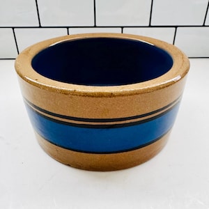 May include: A round, stoneware crock with a wide, tan rim and a dark blue interior. The exterior features a wide blue band with two thin black stripes. The crock is a classic piece of kitchenware.