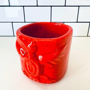 Puede incluir: Una taza de cerámica roja vibrante con una superficie texturizada. La taza presenta un diseño único con detalles en relieve, incluyendo formas circulares y curvas. El interior es de un rojo intenso, y la taza está sobre un fondo blanco.