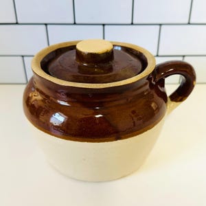 May include: A small, ceramic brown and cream-colored crock pot with a lid and handle. The crock pot has a glossy finish and a light tan rim. The lid has a small, round knob. The handle is curved and attached to the side of the pot.