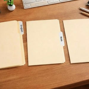 May include: Three manila file folders labeled "English", "Math", and "Art" sit on a wooden desk. The folders are arranged in a row, with the labels visible. A keyboard, pens, and a notebook are in the background.