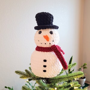 May include: A white crocheted snowman with a black top hat and a red scarf is used as a Christmas tree topper.