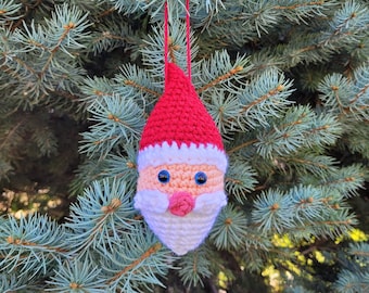 May include: A handmade crochet Santa Claus Christmas ornament. The ornament features a red hat, white beard, and rosy cheeks. Blue eyes and a pink nose complete the festive design. The ornament hangs from a red string, perfect for a Christmas tree.