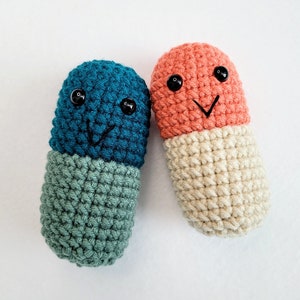 May include: Two crocheted pill-shaped plushies with smiling faces. One is blue and green, the other is orange and cream.