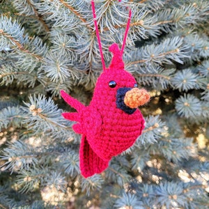 May include: A crocheted cardinal ornament in red with a black beak and orange beak tip. The ornament is hanging from a blue spruce tree.