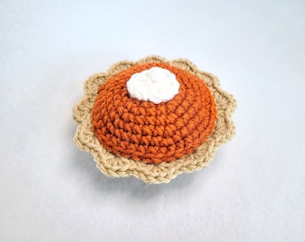 May include: A round, crocheted pumpkin pie ornament featuring a textured, burnt orange filling, a light tan scalloped crust edge, and a small, white, textured dollop of whipped cream centered on top, crafted with visible yarn stitches.