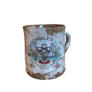 May include: A white ceramic mug with a brown glaze. The mug features a colorful illustration of a castle, a crown, and the text "WALES" and "THE INVESTITURE OF H.R.H. THE PRINCE OF WALES AT CAERNARVON CASTLE JULY 1st 1969".