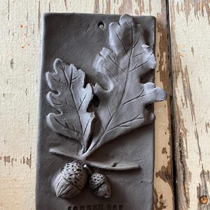 May include: A brown clay tile with a raised design of two oak leaves and two acorns. The tile is signed "COUGER OAK".