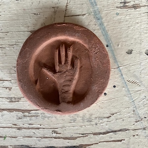 May include: A circular terracotta-colored clay medallion featuring a recessed relief of an open hand with five fingers. The surface of the clay has a matte, earthy texture with visible indentations and a distinct handprint impression centered within the round frame.