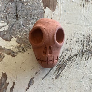 May include: A small, terracotta-colored skull sculpture with large eye sockets and a carved mouth. The skull has a textured surface and is likely handmade. The piece is a decorative item, possibly for Halloween or Day of the Dead.