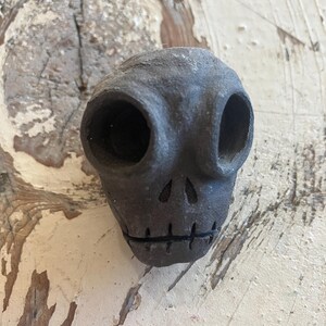 May include: A dark gray ceramic skull with large eye sockets and a carved mouth. The skull has a rough, textured surface and is likely handmade. The design is simple and stylized, suitable for Halloween or gothic decor.