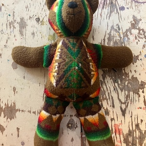May include: A plush bear toy crafted from brown fleece fabric. The bear features a central panel on the torso and legs decorated with a geometric pattern in vibrant shades of green, orange, yellow, and red. It has a black stitched nose and eyes.