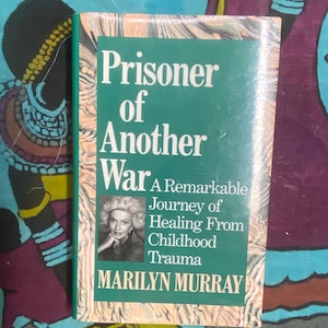 Może przedstawiać: Książka w twardej oprawie zatytułowana "Prisoner of Another War" z zieloną okładką i białym tekstem. Książka zawiera zdjęcie Marilyn Murray i podtytuł "A Remarkable Journey of Healing From Childhood Trauma."