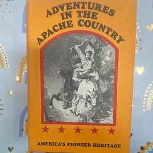 Könnte beinhalten: Ein orangefarbenes Buch mit dem Titel "Adventures in the Apache Country" mit einer Schwarz-Weiß-Illustration von Personen auf Pferden. Das Buch hat einen roten Rand und Sterne mit dem Text "America's Pioneer Heritage" unten.