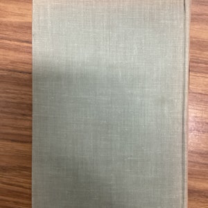 以下が含まれることがあります： 淡い緑色のリネンカバーのハードカバー本。本は閉じられており、テクスチャ加工された表面の裏表紙が表示されています。背表紙は右側に表示されています。本は木製の表面に置かれています。