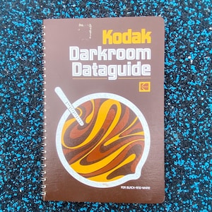 May include: A spiral-bound Kodak Darkroom Dataguide book with a brown cover. The cover features the Kodak logo and the title in yellow and white. The graphic on the cover is a swirl of yellow and brown. The text "FOR BLACK-AND-WHITE" is at the bottom.