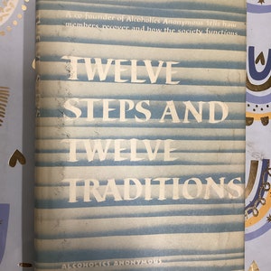 May include: A vintage book titled "Twelve Steps and Twelve Traditions" by Alcoholics Anonymous. The cover is light blue with horizontal stripes and white text. The book's title is prominently displayed, with additional text at the top and bottom.