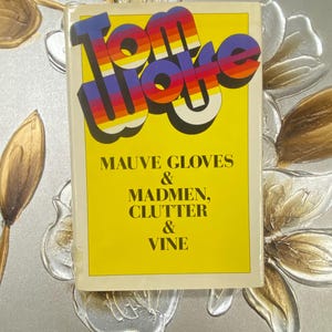 May include: A book titled "Tom Woke" with a yellow cover and retro font. The cover also includes the text "Mauve Gloves & Madmen, Clutter & Vine". The book is set against a floral background.