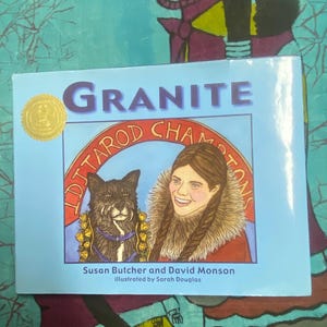 以下が含まれることがあります： 「Granite」というタイトルの子供向けの本で、水色の表紙です。表紙には犬と女性のイラストが描かれており、「Iditarod Champions」というテキストと著者の名前が書かれています。 金色のシールが左上にあります。
