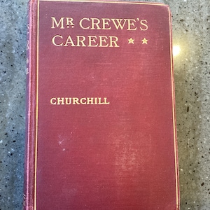 Pode incluir: Um livro encadernado em vinho com letras douradas na capa. O título é "MR. CREWE'S CAREER" com duas estrelas douradas. O nome do autor é "CHURCHILL" em letras douradas.