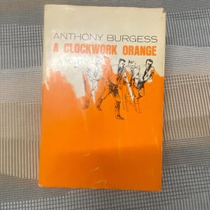 May include: A vintage paperback book titled "A Clockwork Orange" by Anthony Burgess. The cover features an orange and white design with illustrations of figures and the book's title. The book is a novel.